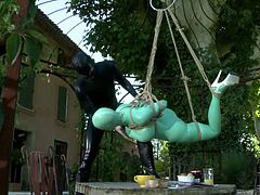 Extremely spoiled babe in latex outfit hangs suspended by ropes in the garden terrace over a table and she is completely at the mercy of her cruel mistress. This hot dominatrix in latex outfit fucks her slave's mouth with her strap-on dildo.